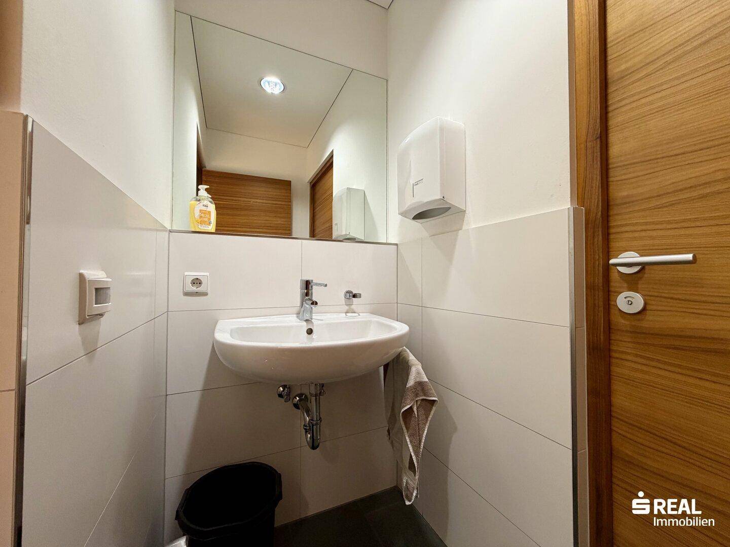 Sauberer Toilettenraum mit Waschbecken, großem Spiegel und weißen Wandfliesen.