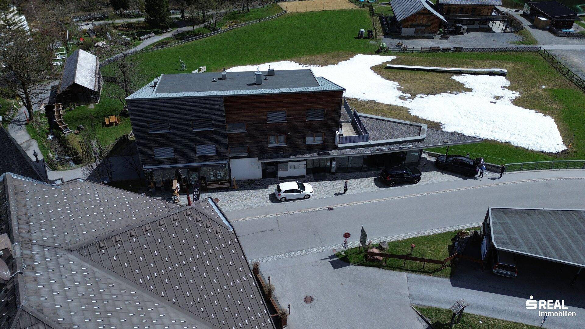 Luftaufnahme des Gebäudes und der umliegenden Landschaft mit Straßen, Grünflächen und vereinzelten Schneeresten.
