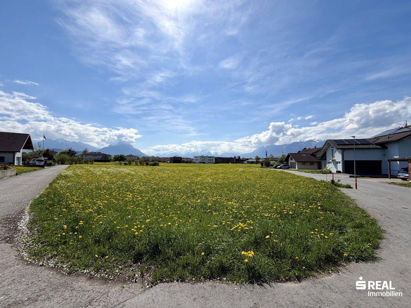 Unbebautes Grundstück mit blühender Wiese unter blauem Himmel, umgeben von Häusern und mit Bergblick.