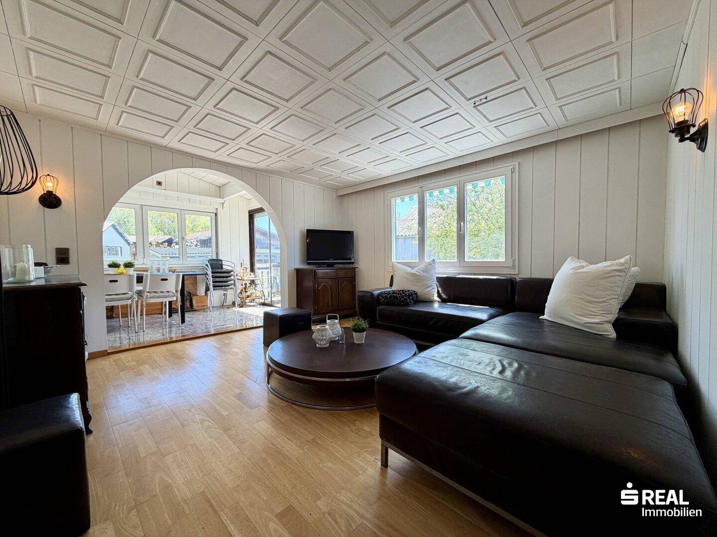 Gemütliches Wohnzimmer mit dunklem Sofa, Holzboden und offenem Zugang zum Essbereich.