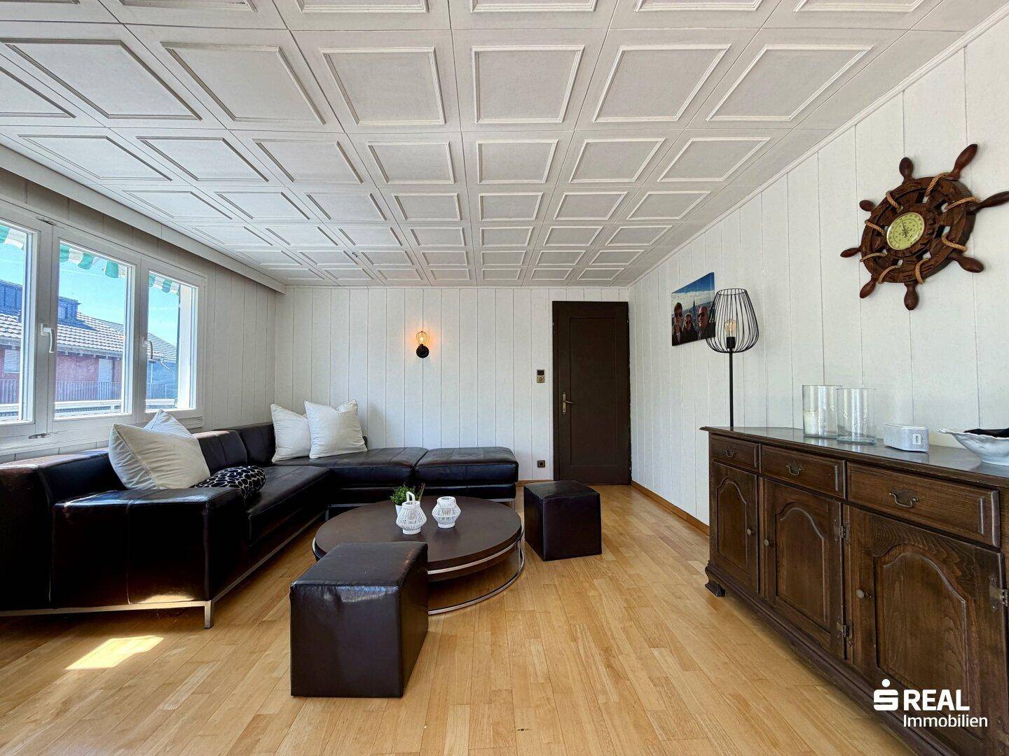 Helles Wohnzimmer mit Holzboden, dunklem Sofa und dekorativer Deckenverkleidung.