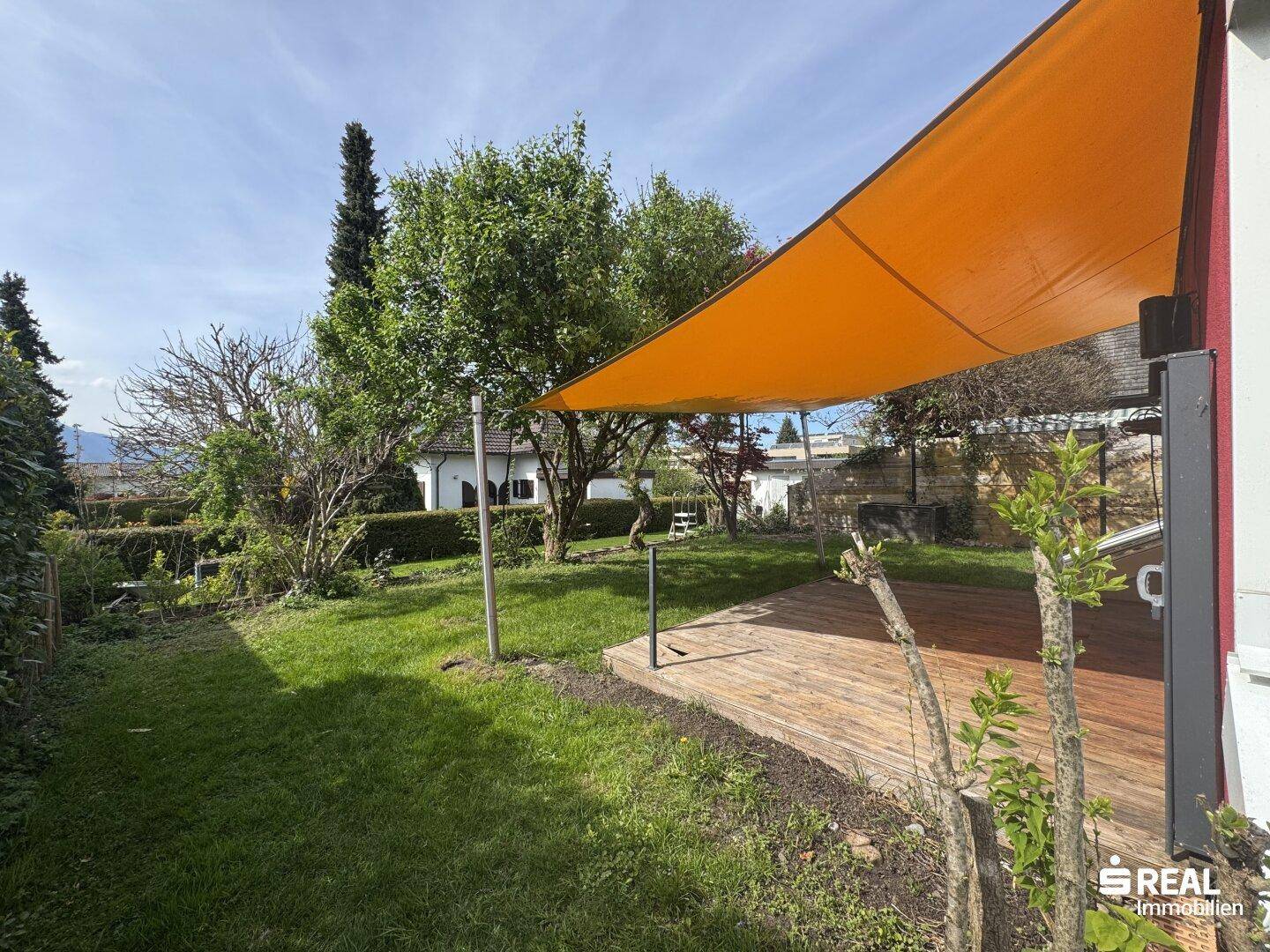 Sonnige Holzterrasse im Garten mit orangefarbenem Sonnensegel und Blick ins Grüne.
