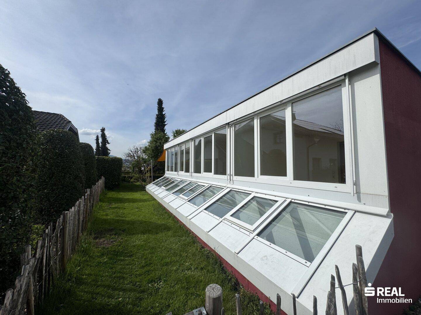 Lange Fensterfront des Gebäudes mit schrägen Fenstern, umgeben von einer Hecke und Rasenfläche.