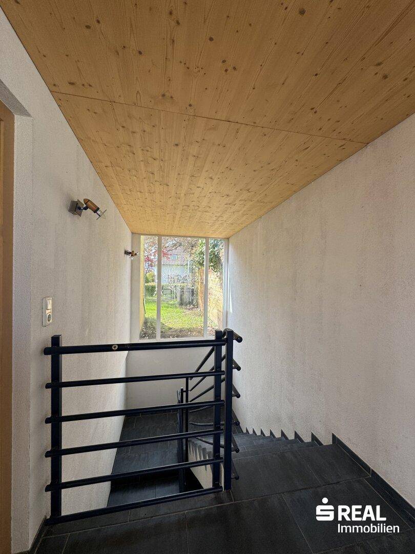 Treppenhaus mit Holzdecke und Blick in den Garten durch ein großes Fenster.