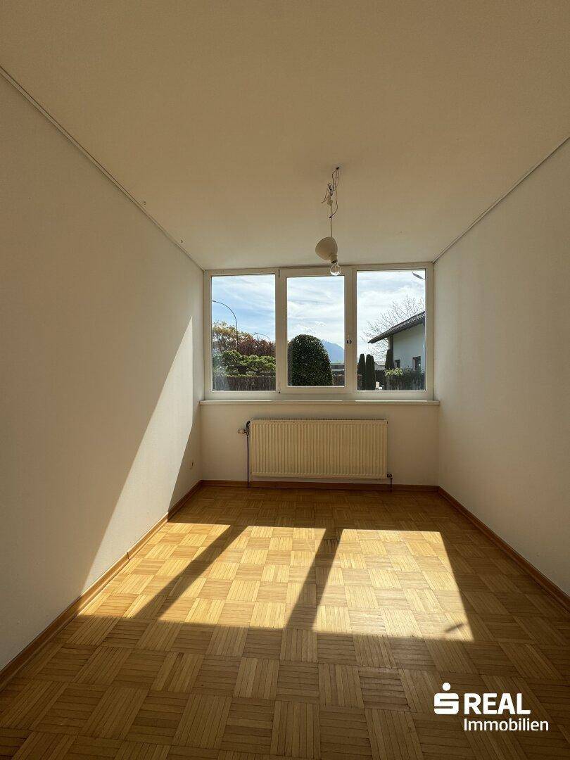 Sonniger Raum mit Parkettboden und großem Fenster, das einen Blick ins Grüne bietet.