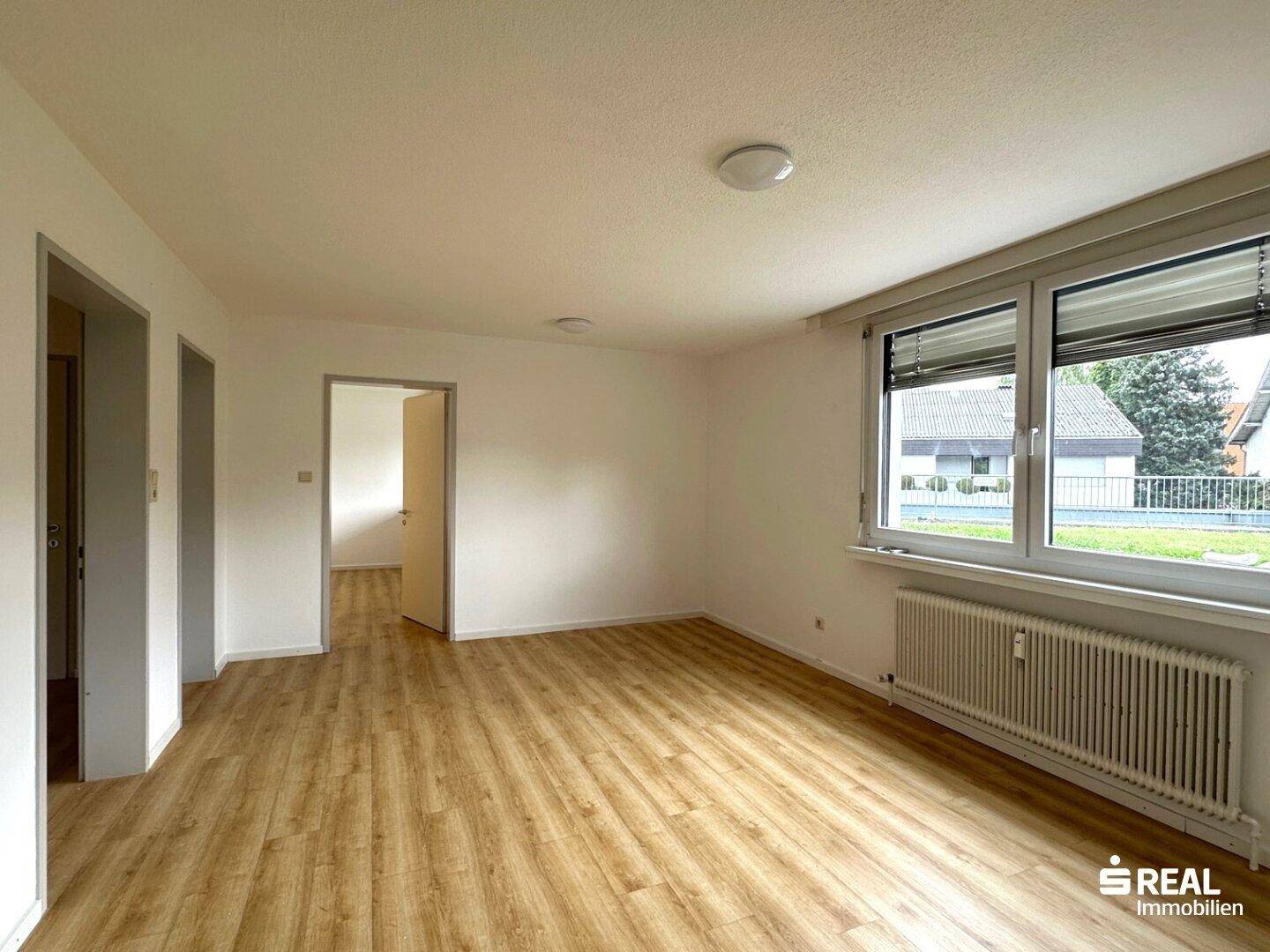 Heller, leerer Raum mit Laminatboden und großen Fenstern, die einen Blick nach draußen bieten.