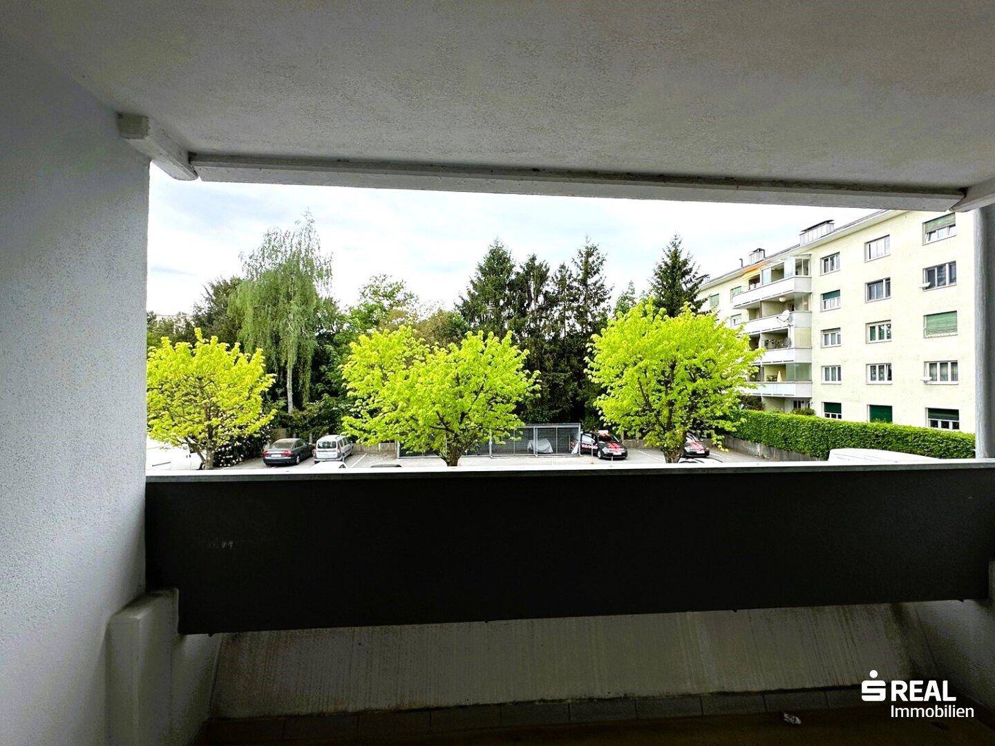 Balkon mit Blick auf grüne Bäume und einen Parkplatz, umgeben von Wohngebäuden.