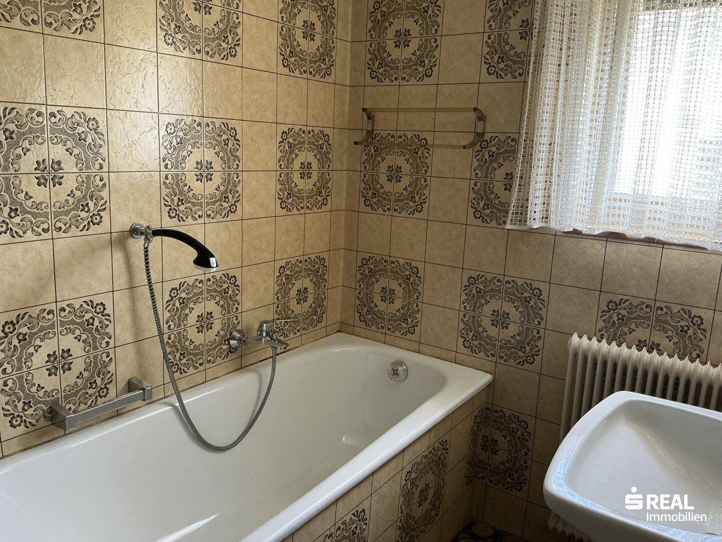 Badezimmer mit beigefarbenen Fliesen, Badewanne und Fenster für Tageslicht.