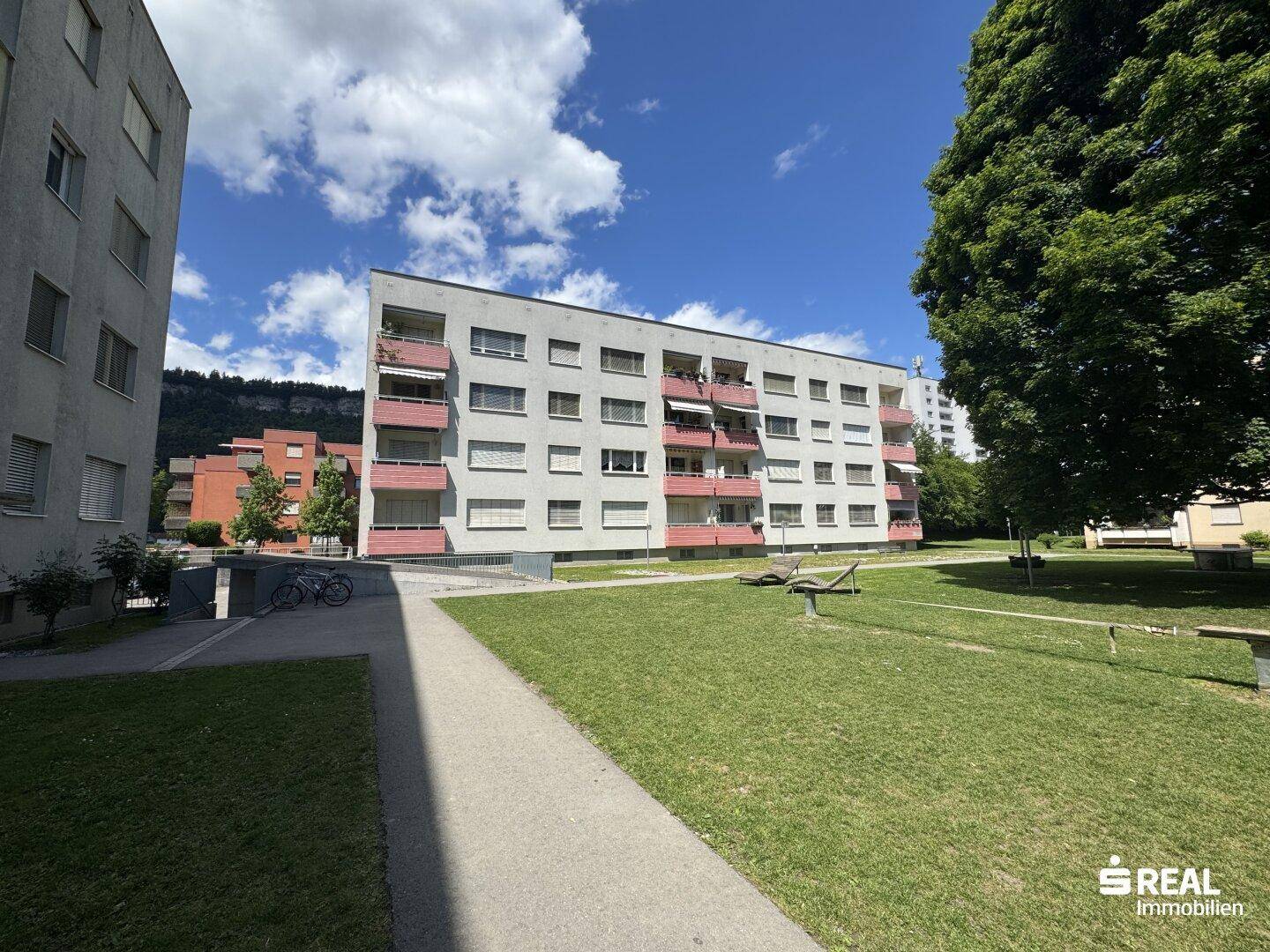 Wohngebäude mit Balkonen, umgeben von Grünflächen und Bäumen, mit Bergblick.