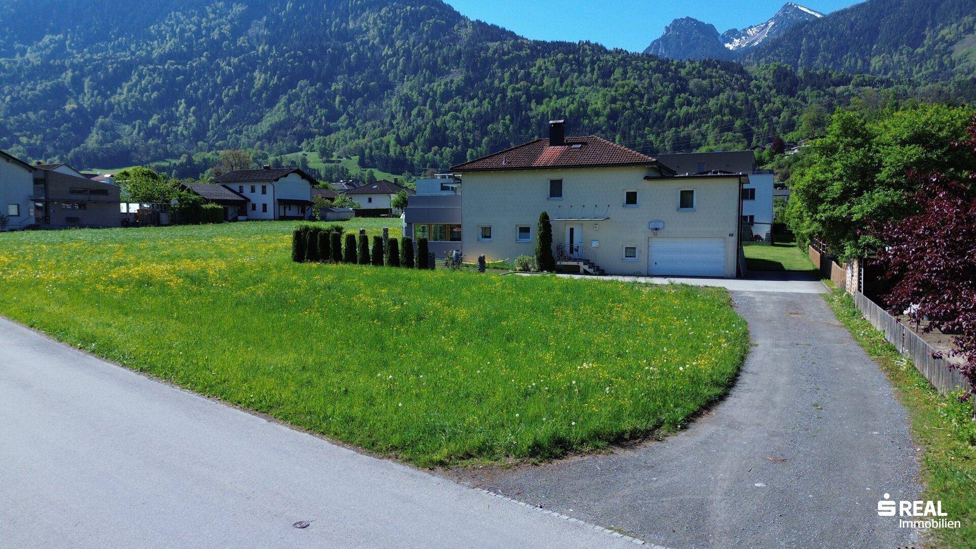 Sonniges Baugrundstück mit grüner Wiese, angrenzendem Haus und beeindruckender Bergkulisse.