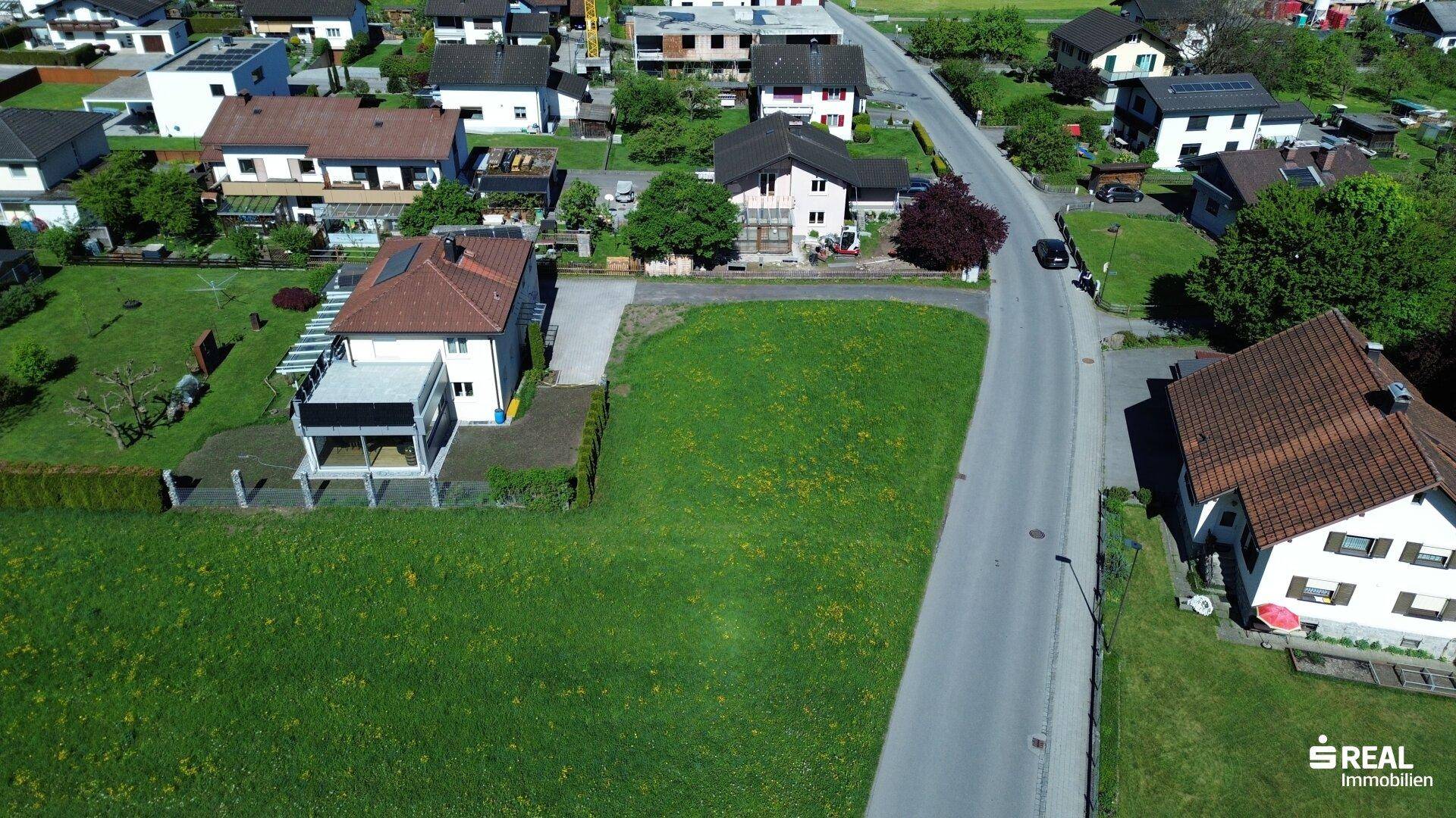 Luftaufnahme des grünen Baugrundstücks in einer Wohnsiedlung mit angrenzender Straße und Häusern.