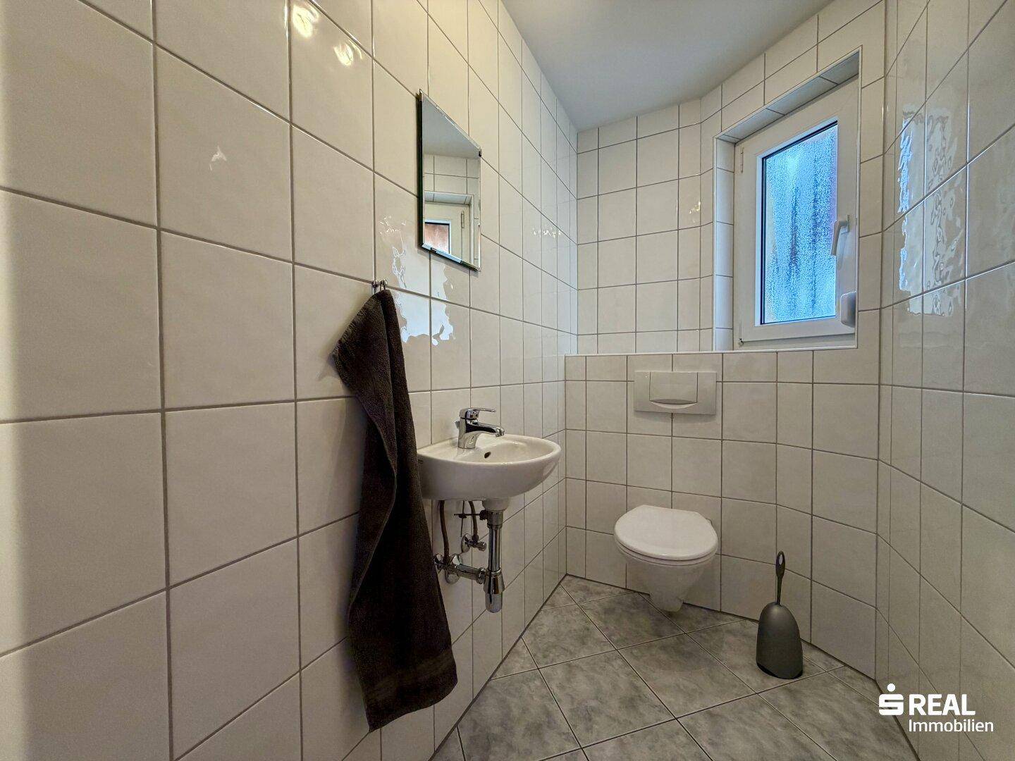 Kleines Gäste-WC mit weißen Fliesen, wandhängendem WC und einem Fenster für Tageslicht.