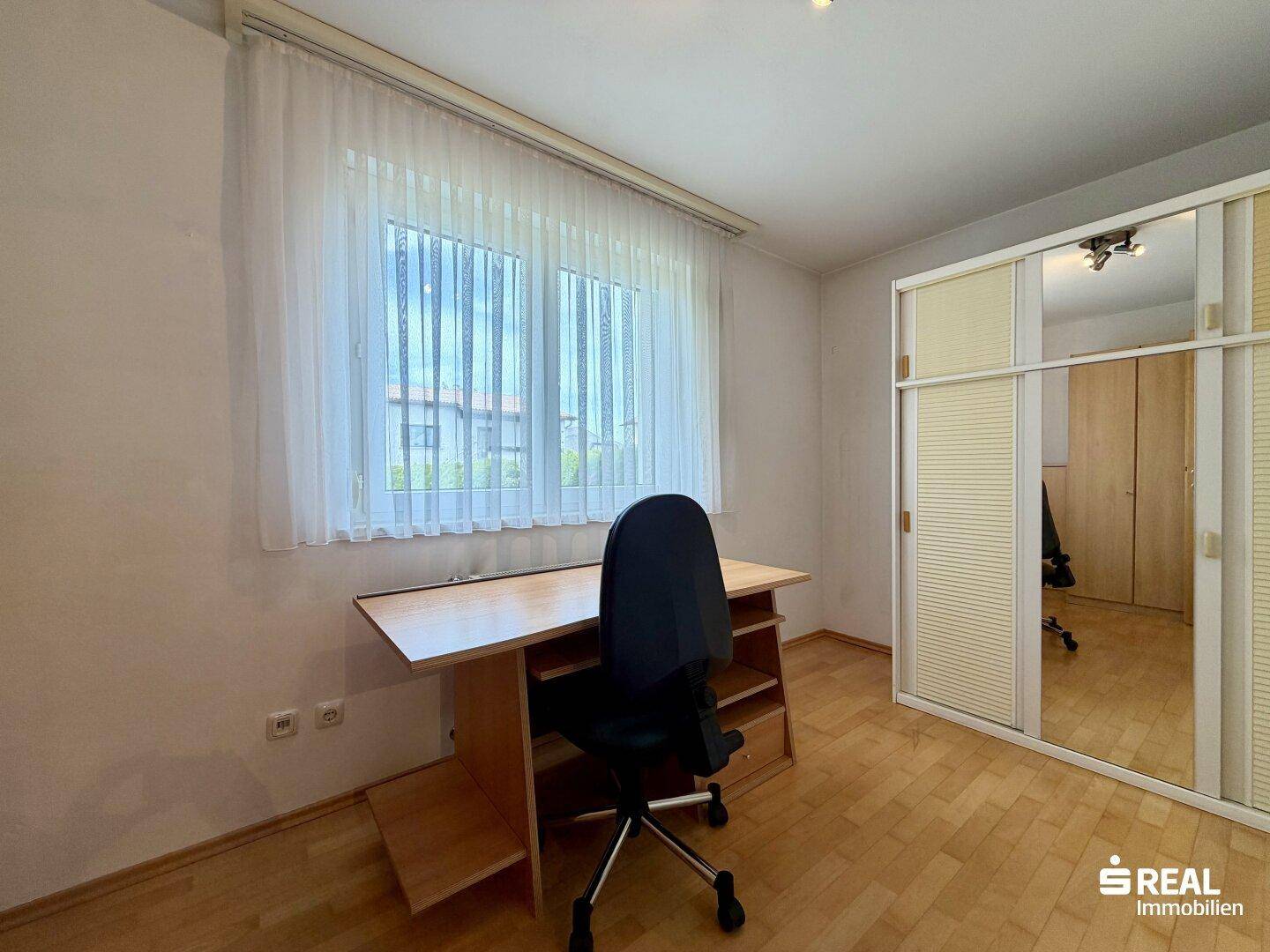Helles Arbeitszimmer mit Parkettboden, großem Fenster und einem Kleiderschrank mit Spiegeltüren.