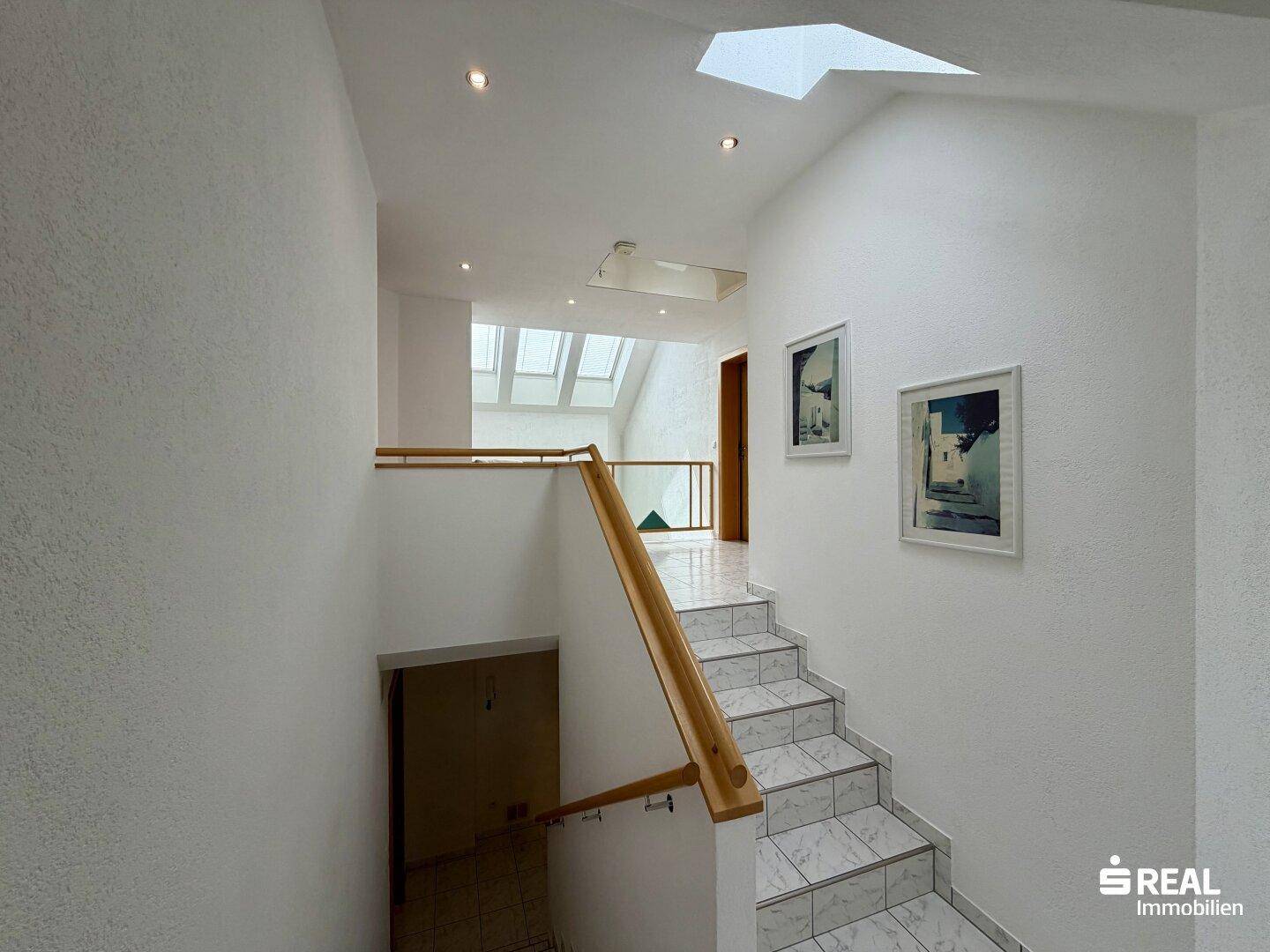 Treppenhaus mit Fliesenstufen, Holzgeländer und Blick auf die obere Galerie mit Dachfenstern.