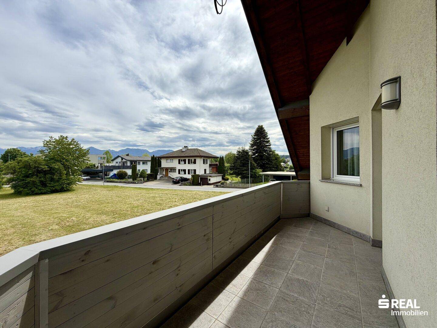 Balkon mit Holzboden und Geländer, bietet einen weiten Blick über die grüne Umgebung und Nachbarschaft.