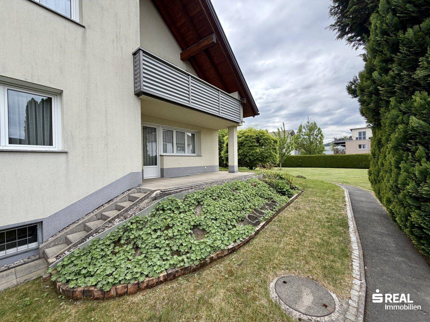 Außenbereich des Hauses mit kleiner Terrasse, Treppe und bepflanztem Gartenbeet entlang des Weges.