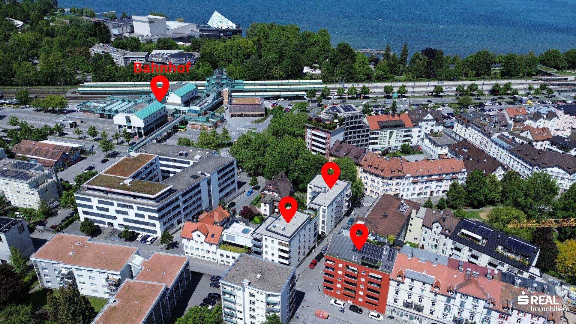 Stadtansicht aus der Vogelperspektive mit See, Bahnhof und umliegenden Gebäuden unter blauem Himmel.