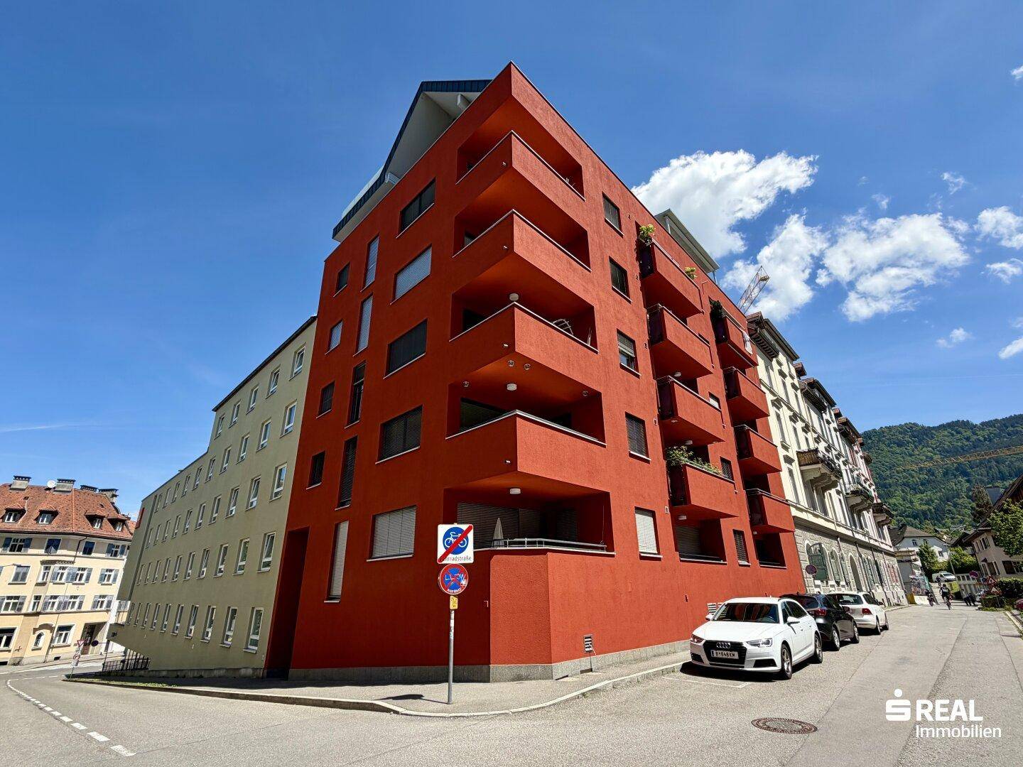 Moderne Wohnfassade in leuchtendem Rot mit Balkonen unter blauem Himmel und Bergen im Hintergrund.