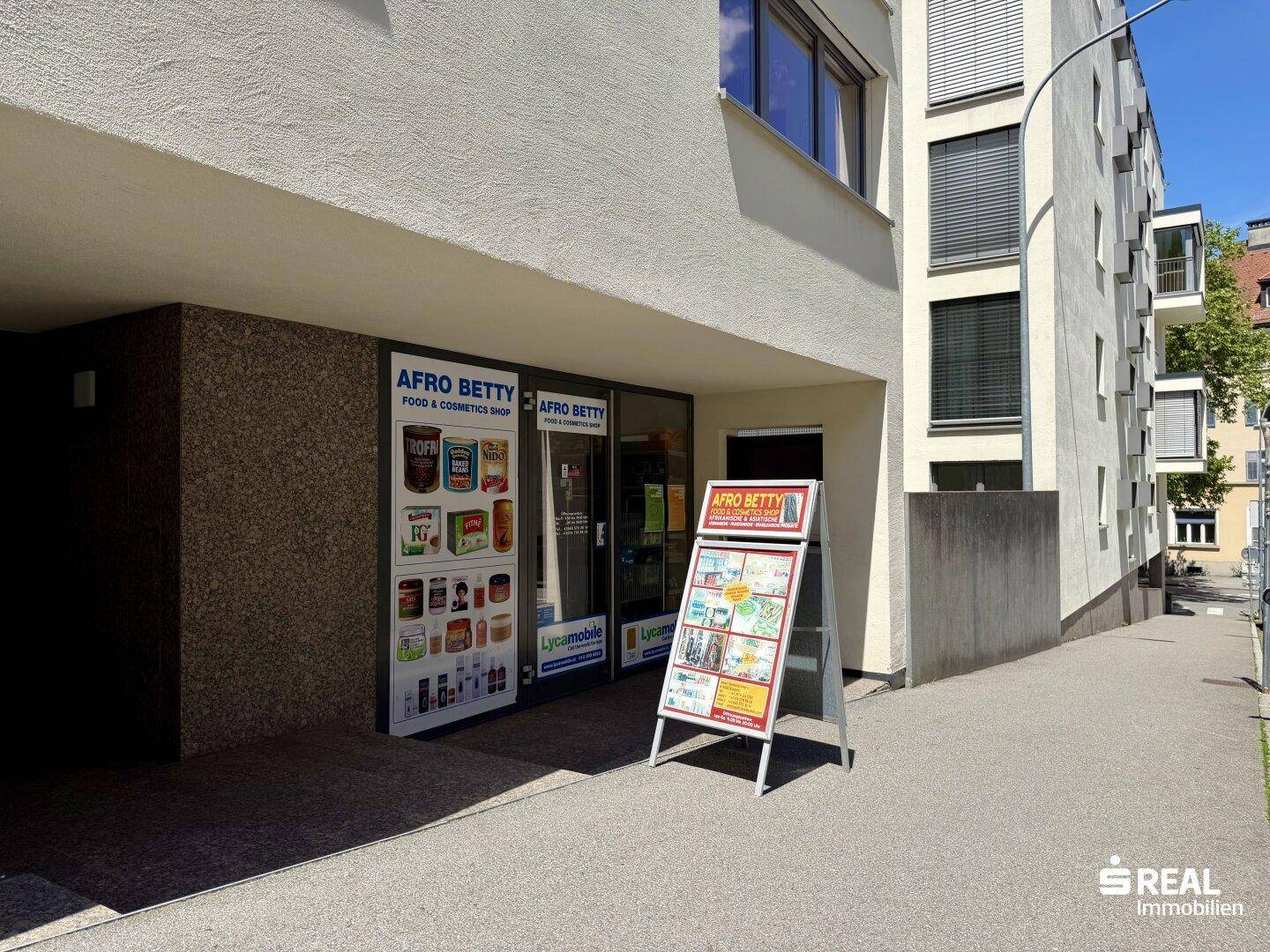 Eingangsbereich eines Geschäfts im Erdgeschoss mit Werbetafeln und Schaufenstern an einem sonnigen Tag.