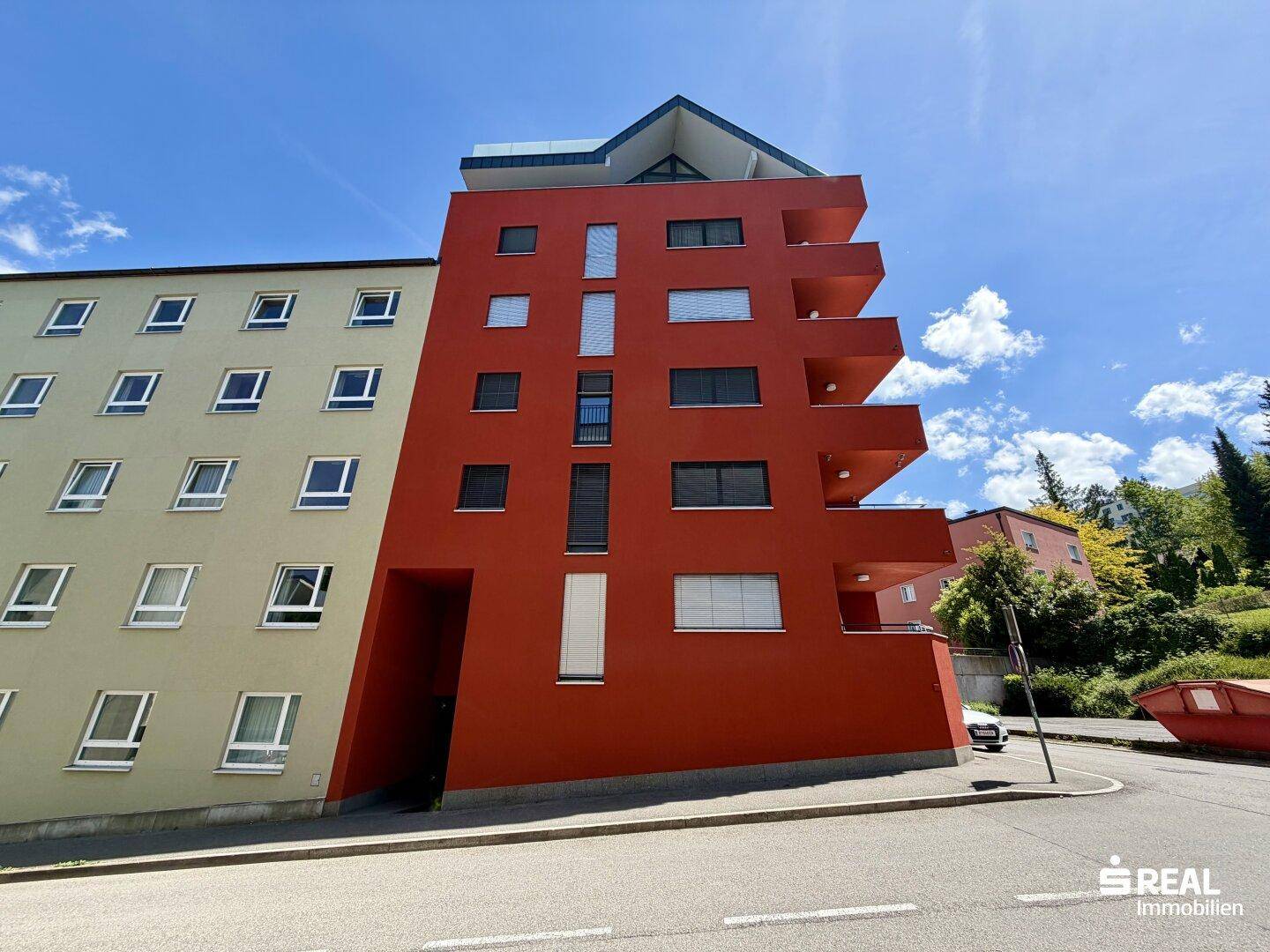 Rote Gebäudefassade mit vertikalen Fenstern und angrenzendem hellen Gebäude, umgeben von Bäumen.