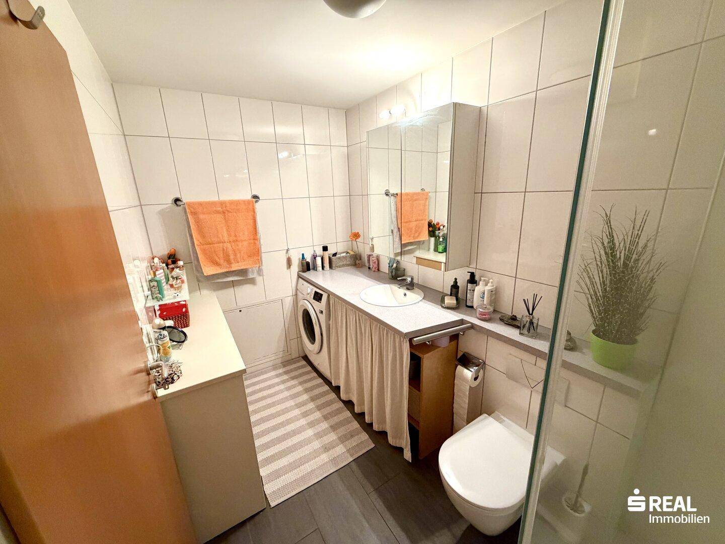 Badezimmer mit Duschkabine, Waschbecken und integrierter Waschmaschine, gefliest in Weiß und Dunkelgrau.