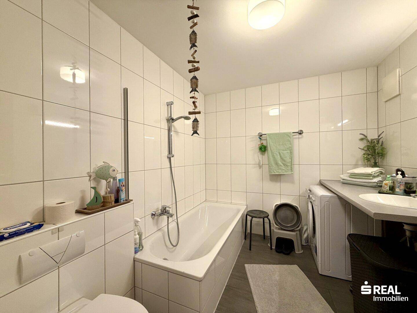 Badezimmer mit Badewanne, Waschbecken und WC, gefliest in Weiß und Dunkelgrau, inklusive Waschmaschine.