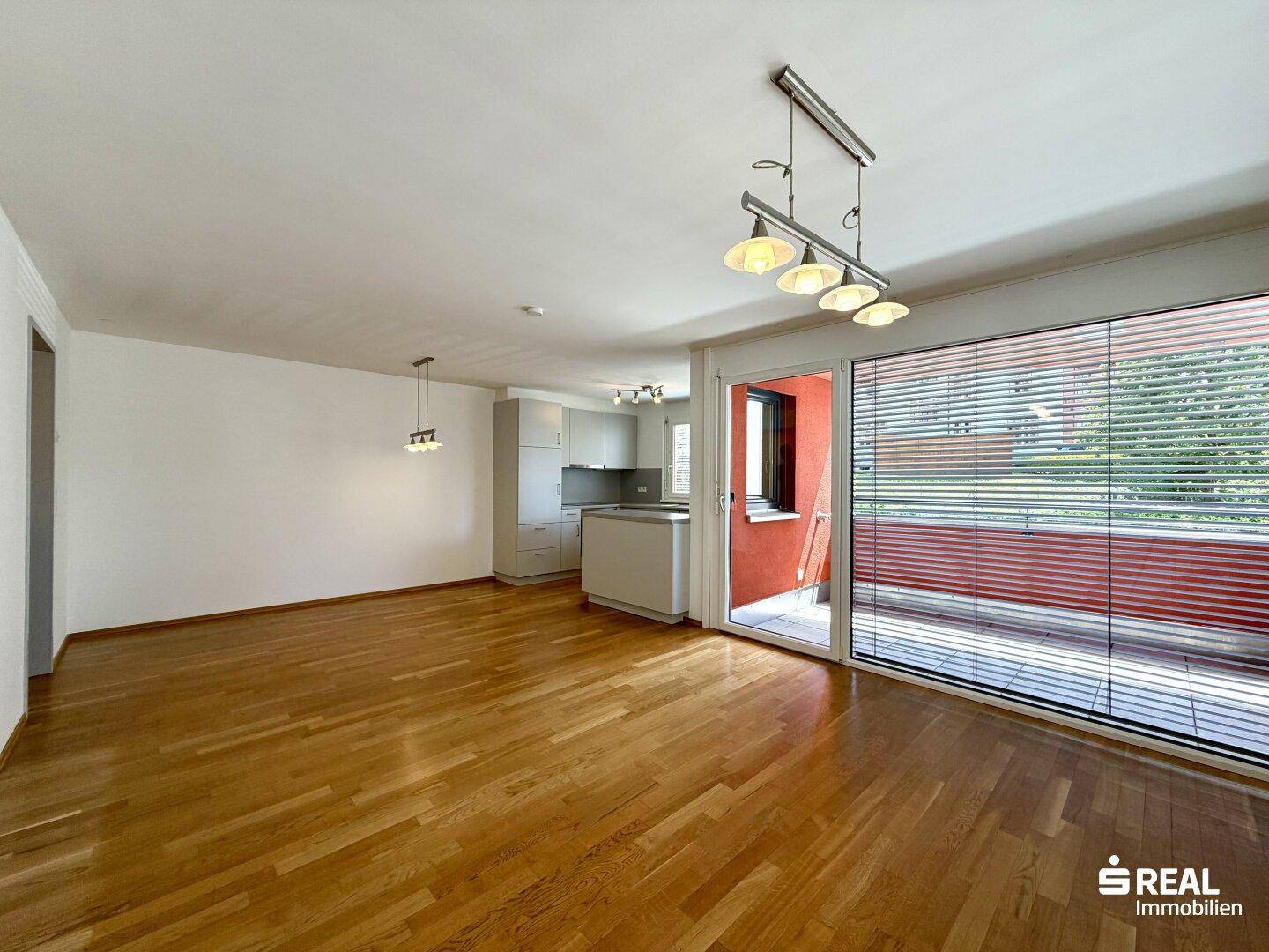 Geräumiger offener Wohnbereich mit Holzboden und integrierter Küche, großer Fensterfront und Balkonzugang.