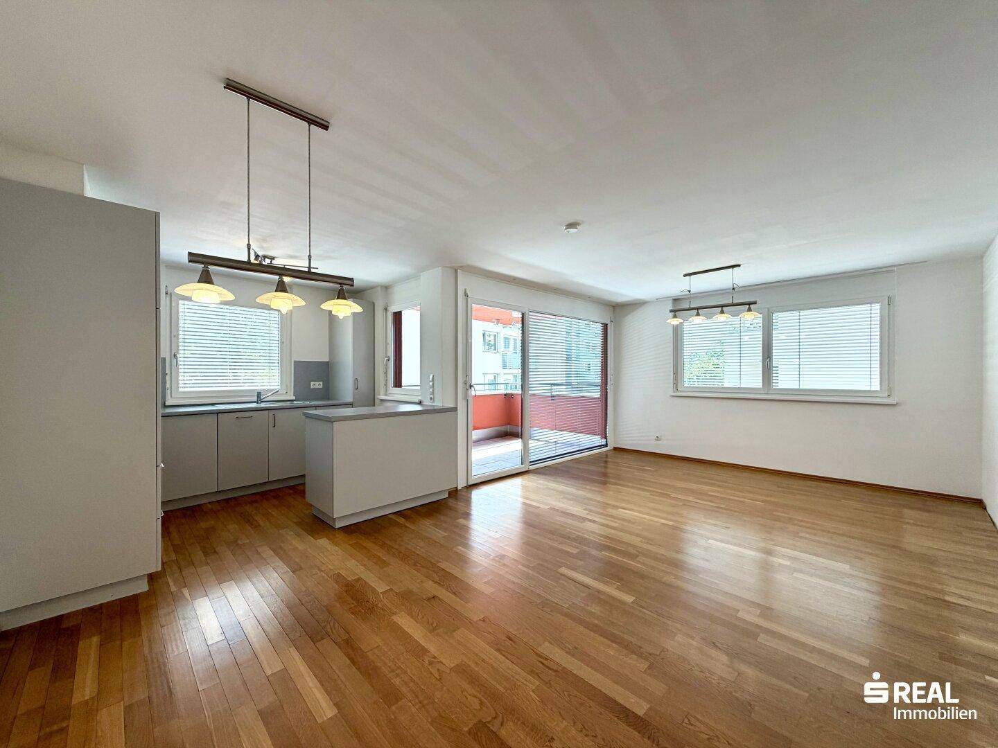Heller offener Wohnbereich mit Holzboden und integrierter Küche, Zugang zum Balkon und viel Tageslicht.
