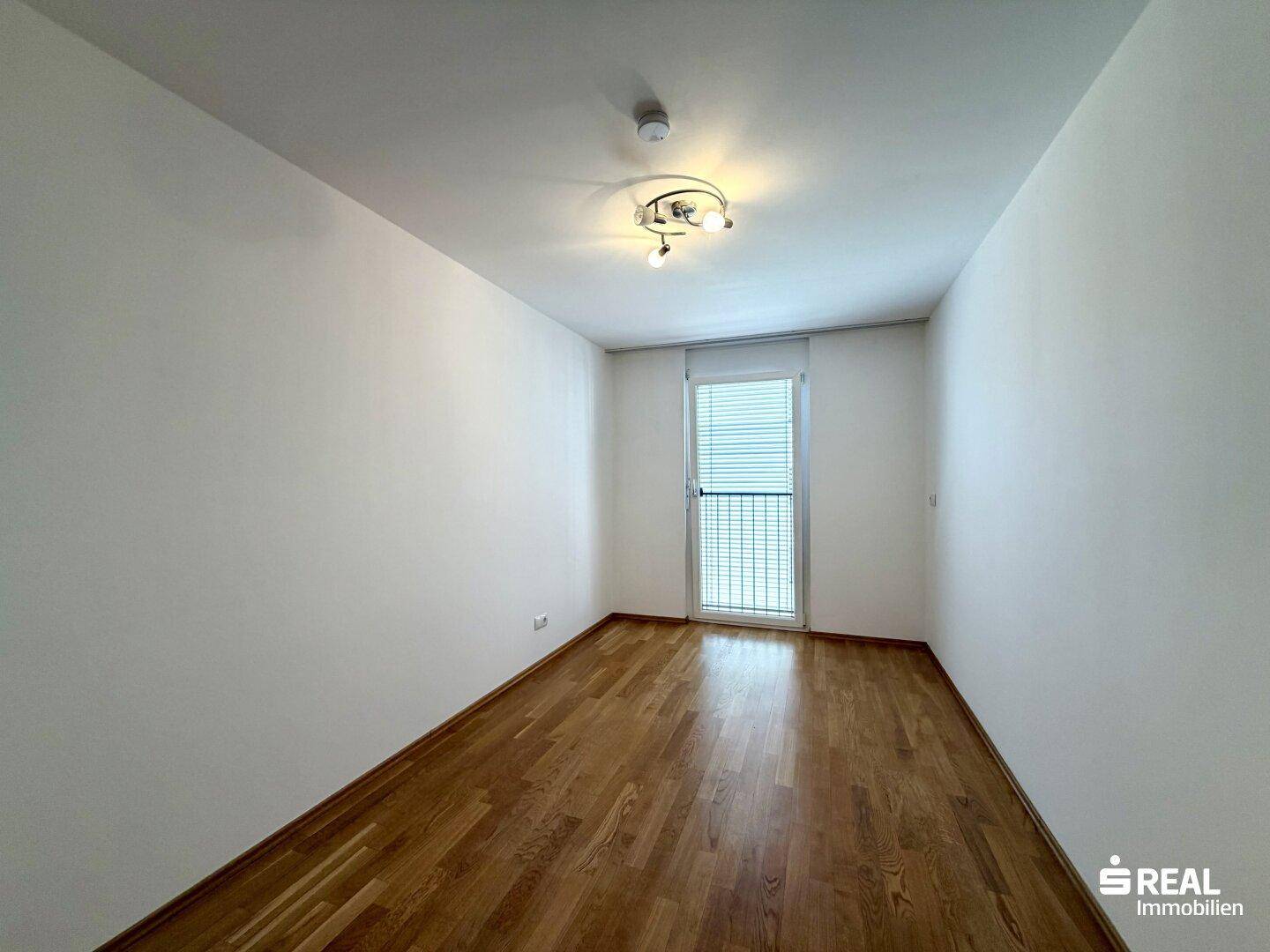 Leeres Schlafzimmer mit Holzboden, großem Fenster und viel Tageslicht, bereit zur Einrichtung.