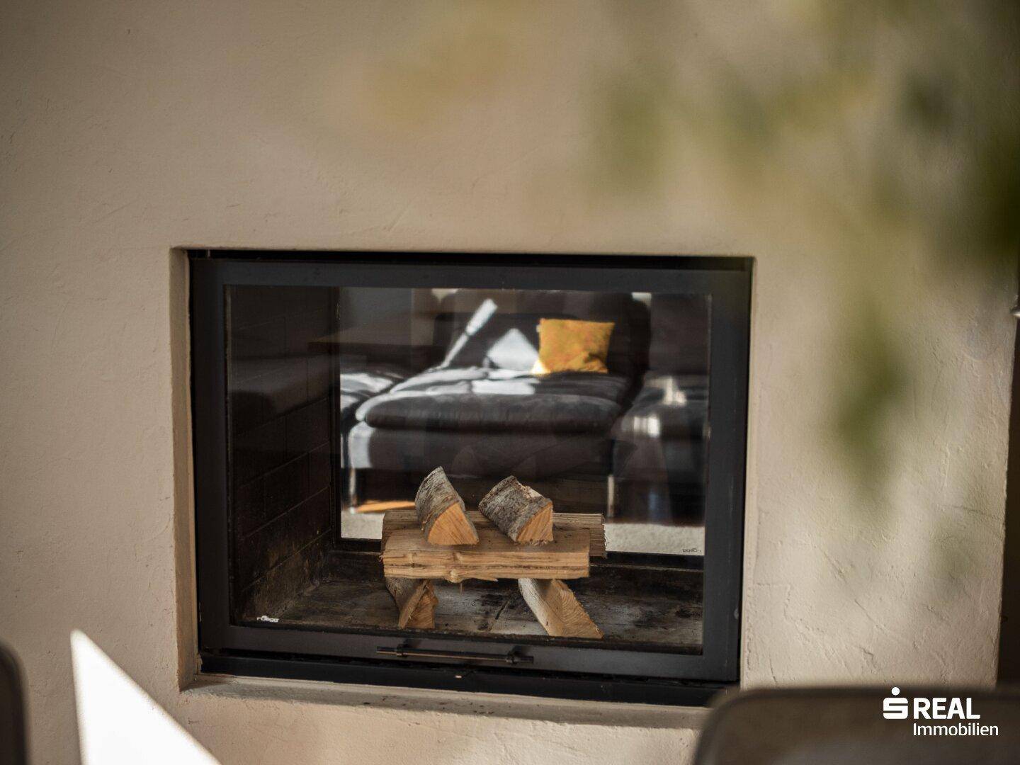 Kamin mit Holzscheiten und Spiegelung des gemütlichen Wohnbereichs im Glas.