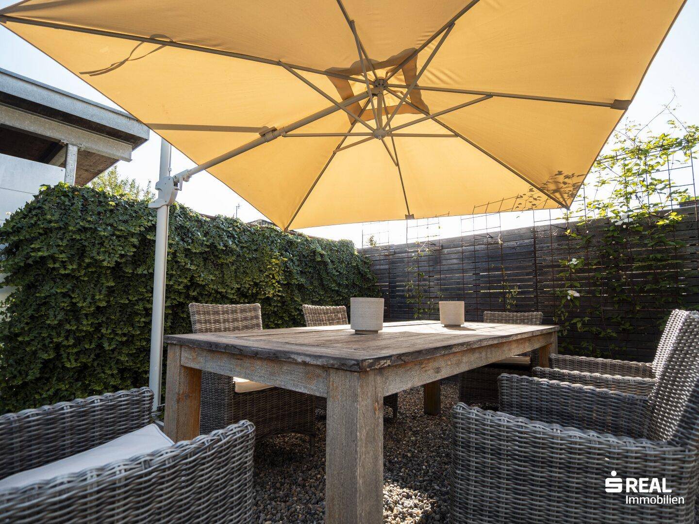 Gemütlicher Essbereich im Garten mit großem Holztisch und Sonnenschirm.