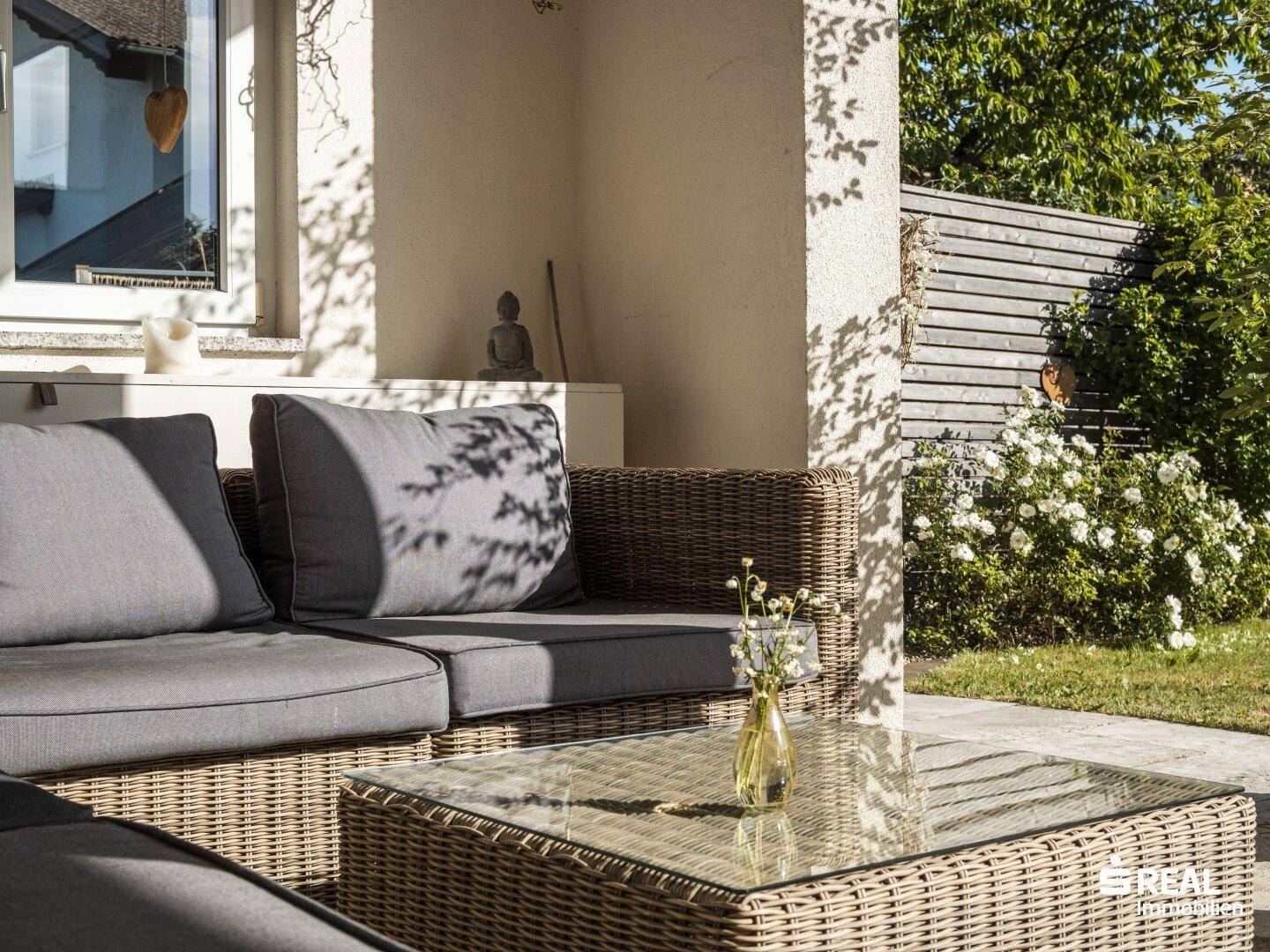 Gemütliche Sitzecke im Garten mit Rattanmöbeln und blühenden Pflanzen.