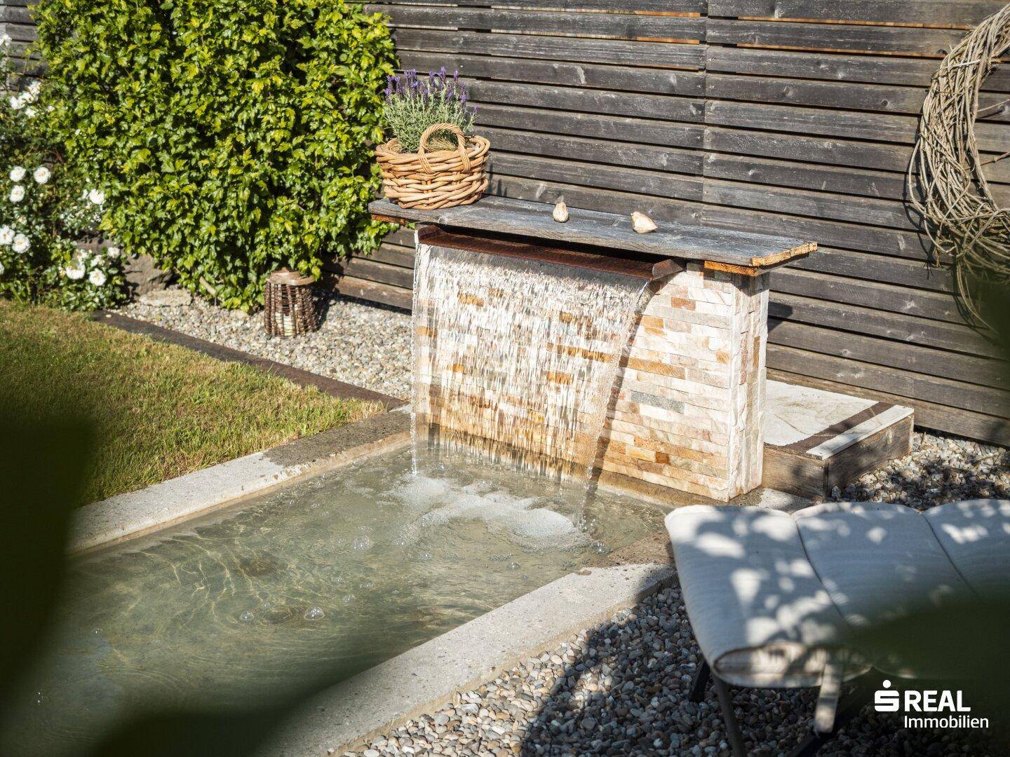 Nahaufnahme des Wasserfalls im Garten, umgeben von Kies und Grün.