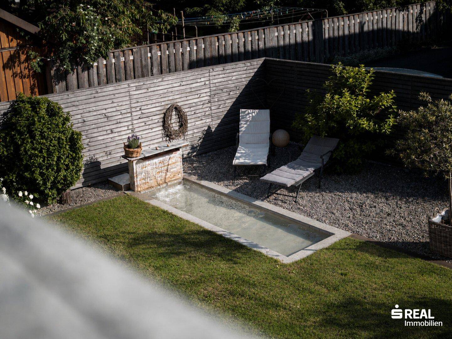 Gepflegter Gartenbereich mit kleinem Wasserbecken, Liegestühlen und Holzzaun.
