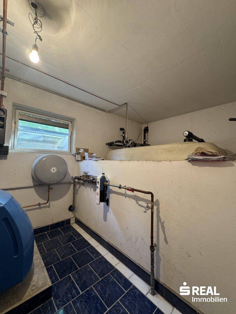 Technikraum im Keller mit Heizungsinstallationen, Öltank und dunklen Fliesen.