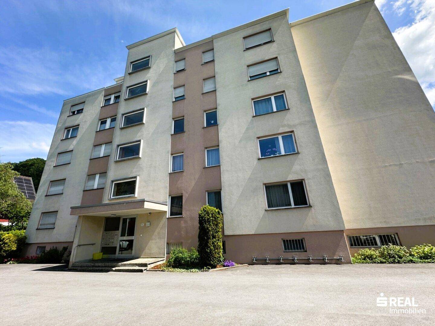 Mehrstöckiges Apartmentgebäude mit heller Fassade und einem Vorplatz, der als Parkbereich dient.