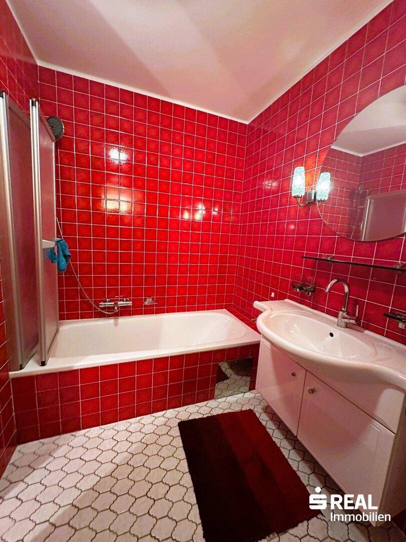 Badezimmer mit leuchtend roten Fliesen, einer Badewanne mit Duschabtrennung und einem Waschbecken mit Spiegel.
