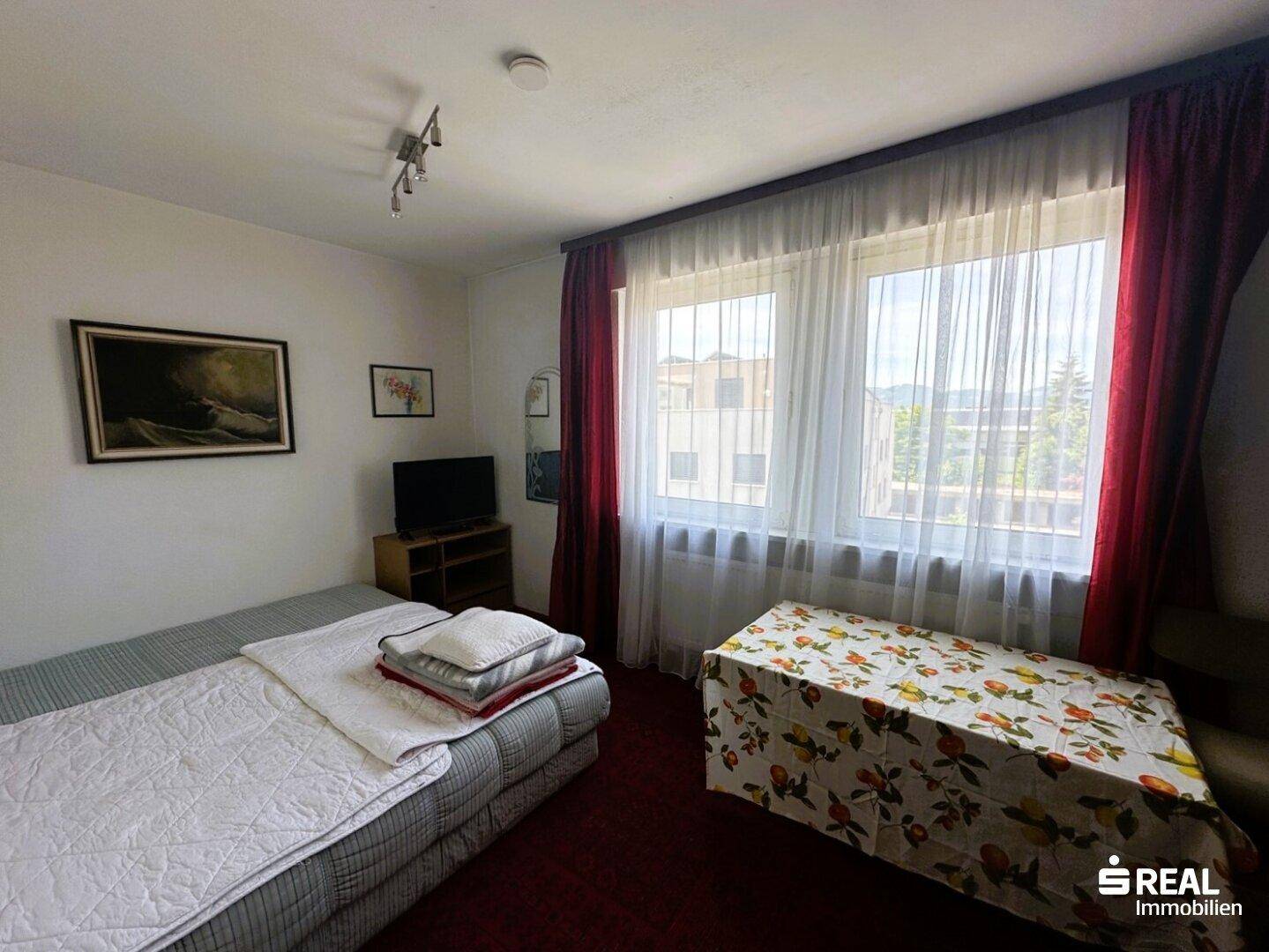 Schlafzimmer mit Einzelbett, kleinem Tisch und Fenster, ausgestattet mit einem roten Teppichboden.