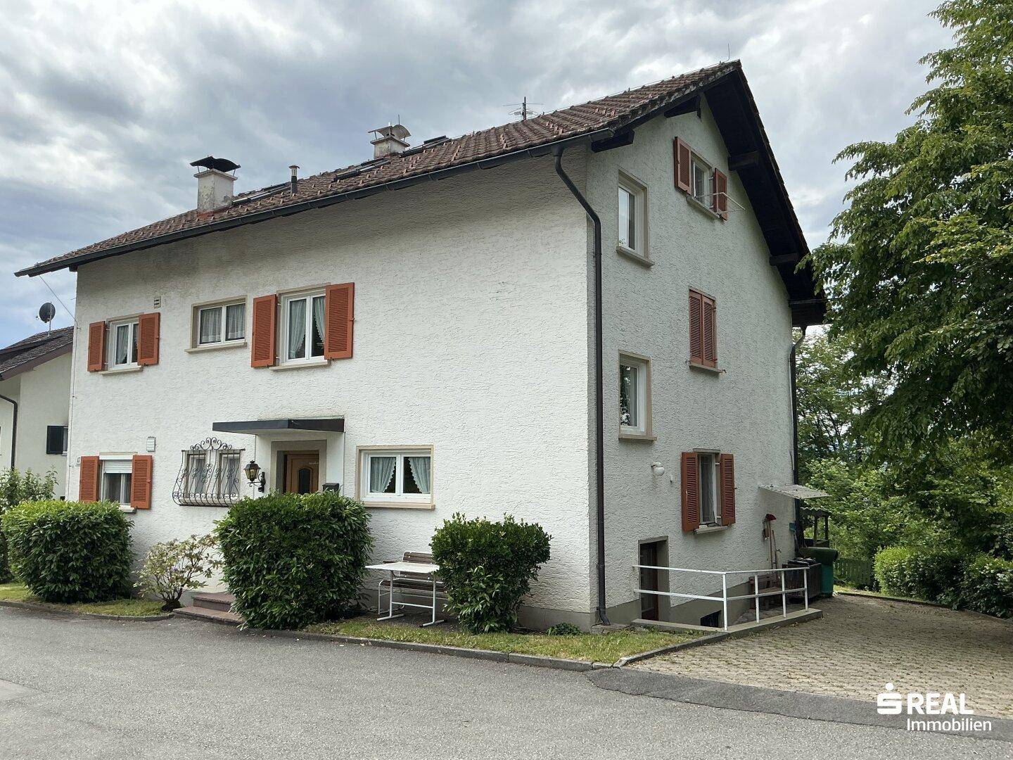 Seitenansicht des weißen Hauses mit roten Fensterläden, umgeben von Bäumen und Sträuchern.