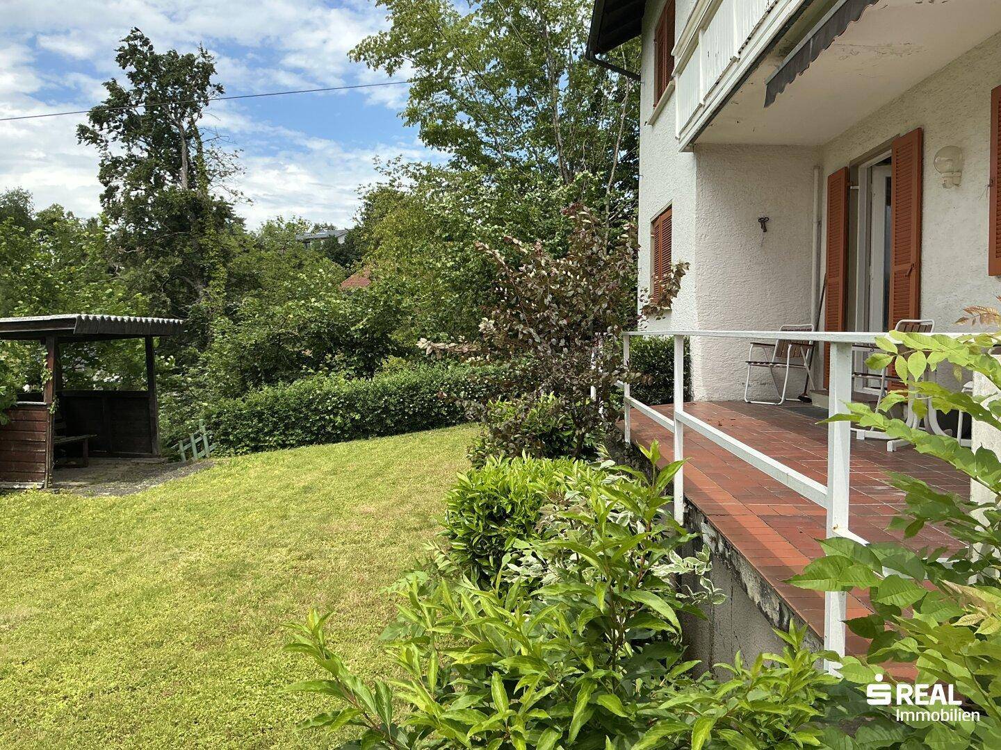 Grüner Gartenbereich mit Rasenfläche, Hecken und einer kleinen überdachten Sitzgelegenheit im Freien.