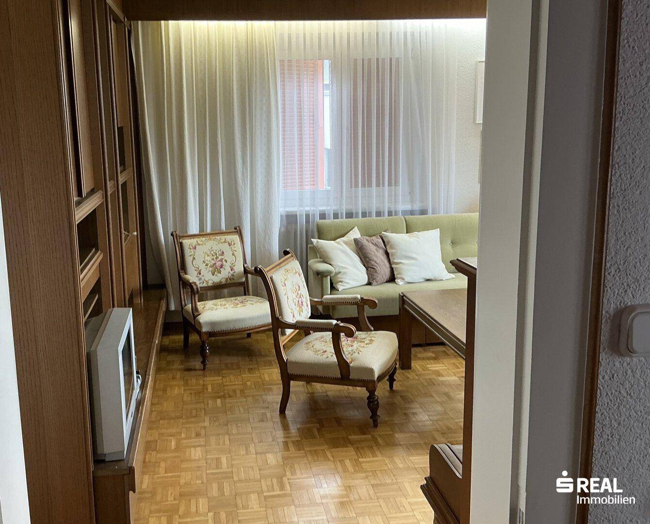 Wohnzimmer mit Holzvertäfelung, traditionellen Sesseln und einem Sofa, durch Fenster gut beleuchtet.