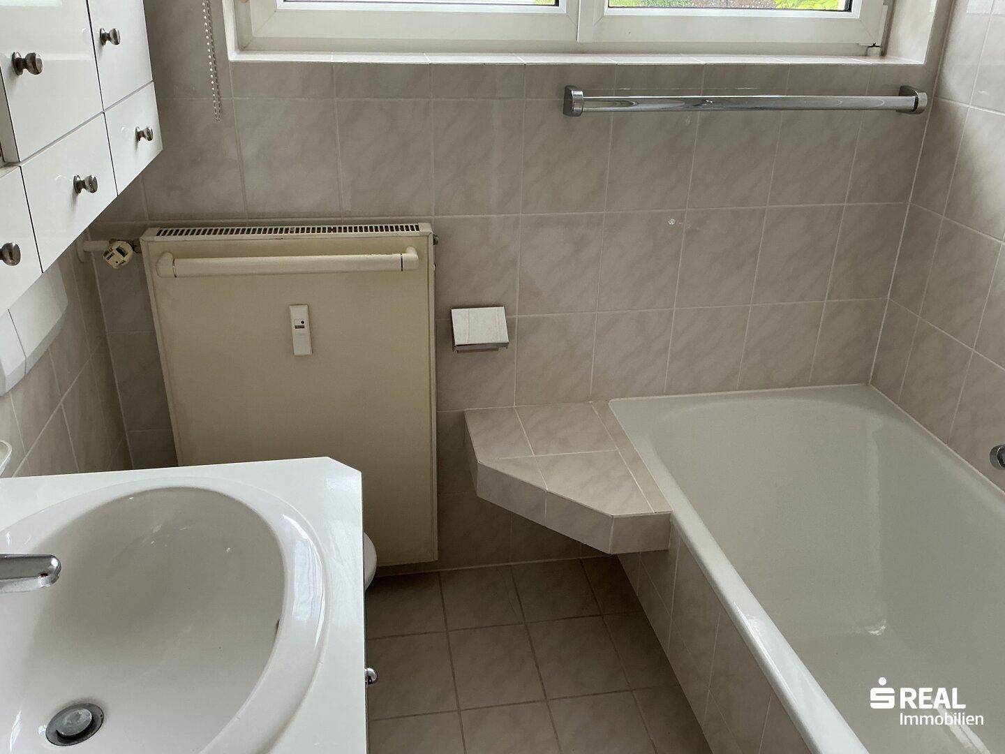 Badezimmer mit Badewanne, Waschbecken und hellen Fliesen, ausgestattet mit einem Fenster.