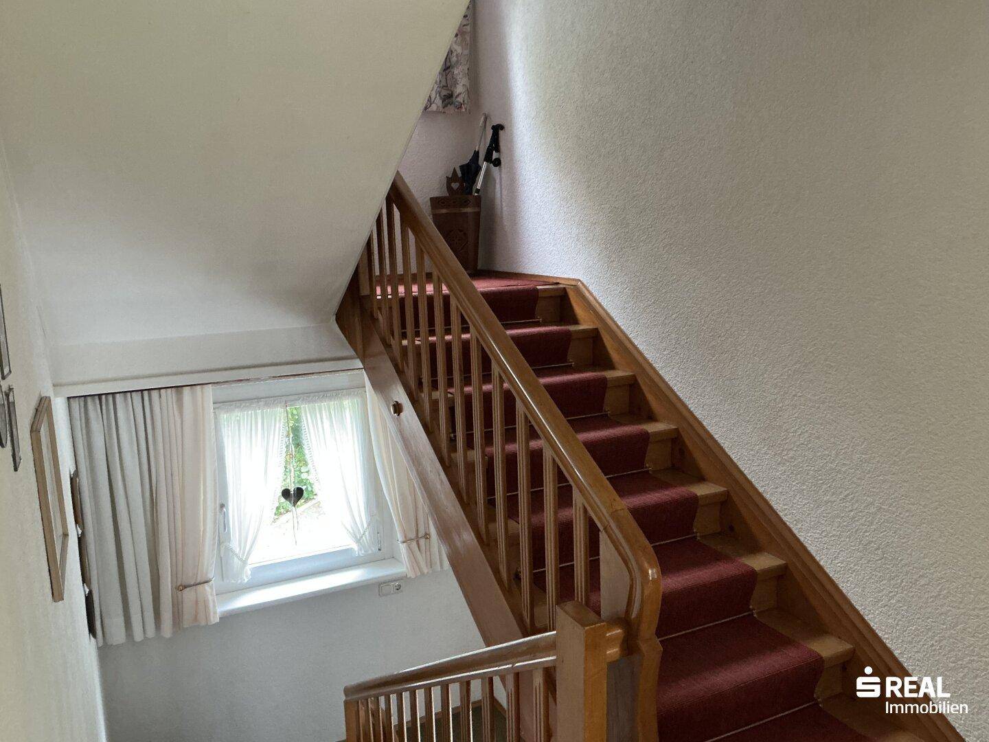 Holztreppe mit rotem Teppichläufer führt in die obere Etage, beleuchtet durch ein Fenster.