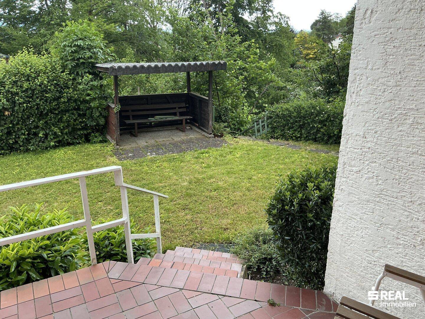 Gartenbereich mit Rasenfläche, Treppe und einer überdachten Sitzgelegenheit im Hintergrund.