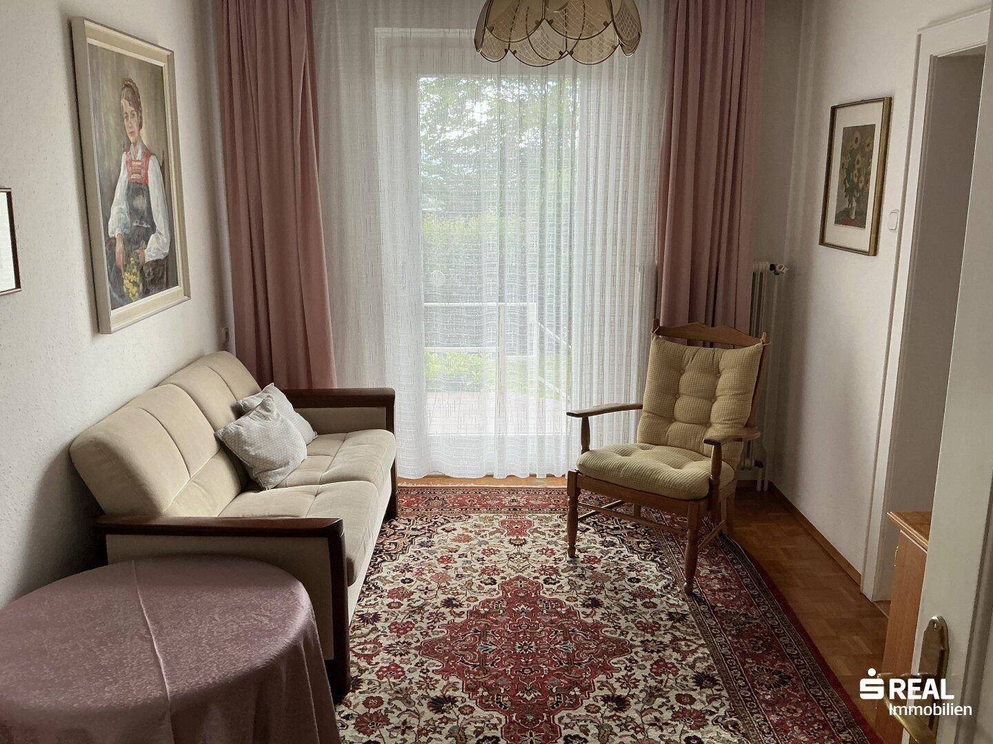 Gemütliches Schlafzimmer mit Sofa, Sessel und Zugang zu einem Balkon, hell durch die Tür.