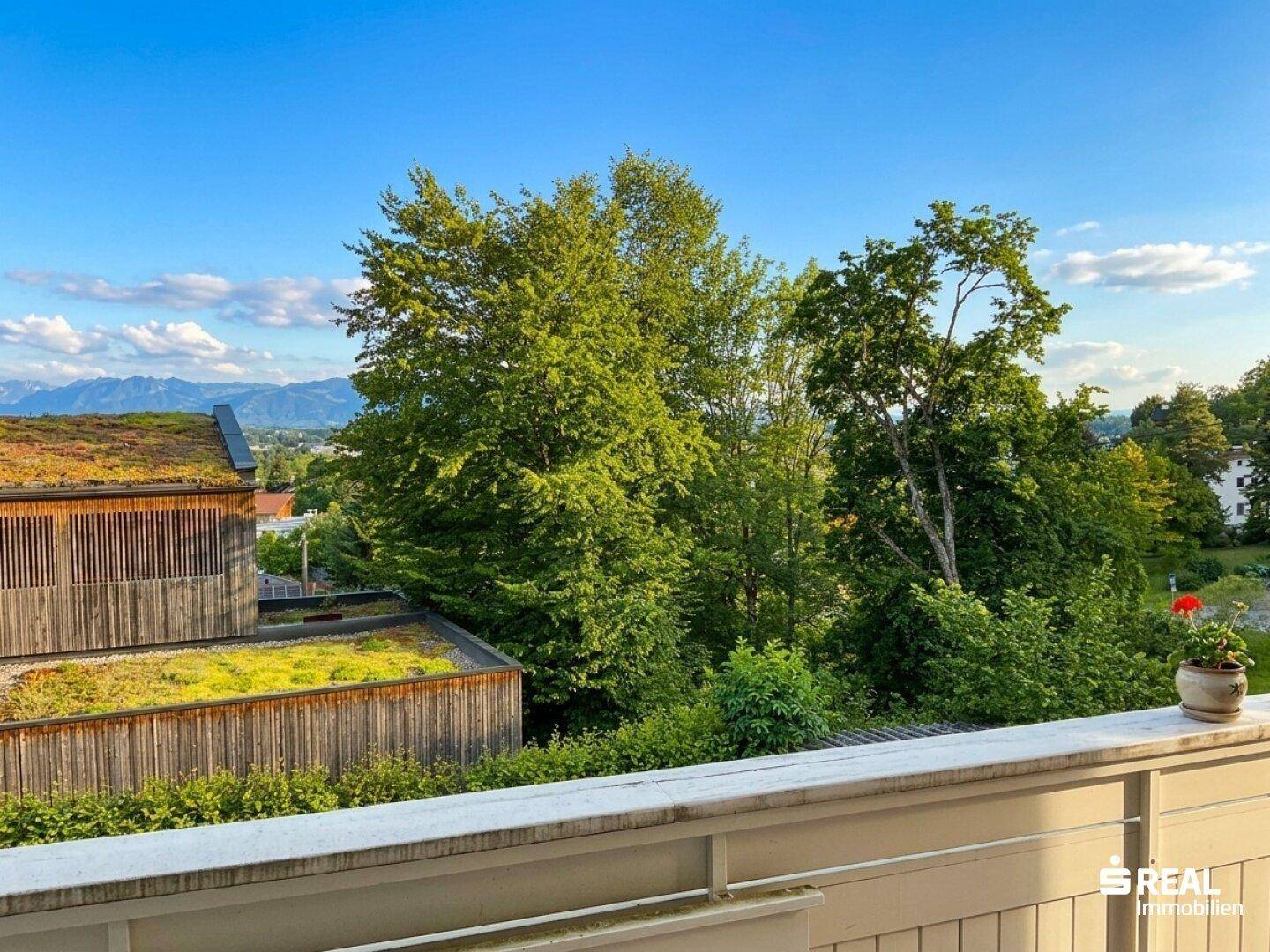 Panoramablick vom Balkon auf grüne Bäume, Dächer mit Begrünung und ferne Berge unter blauem Himmel.