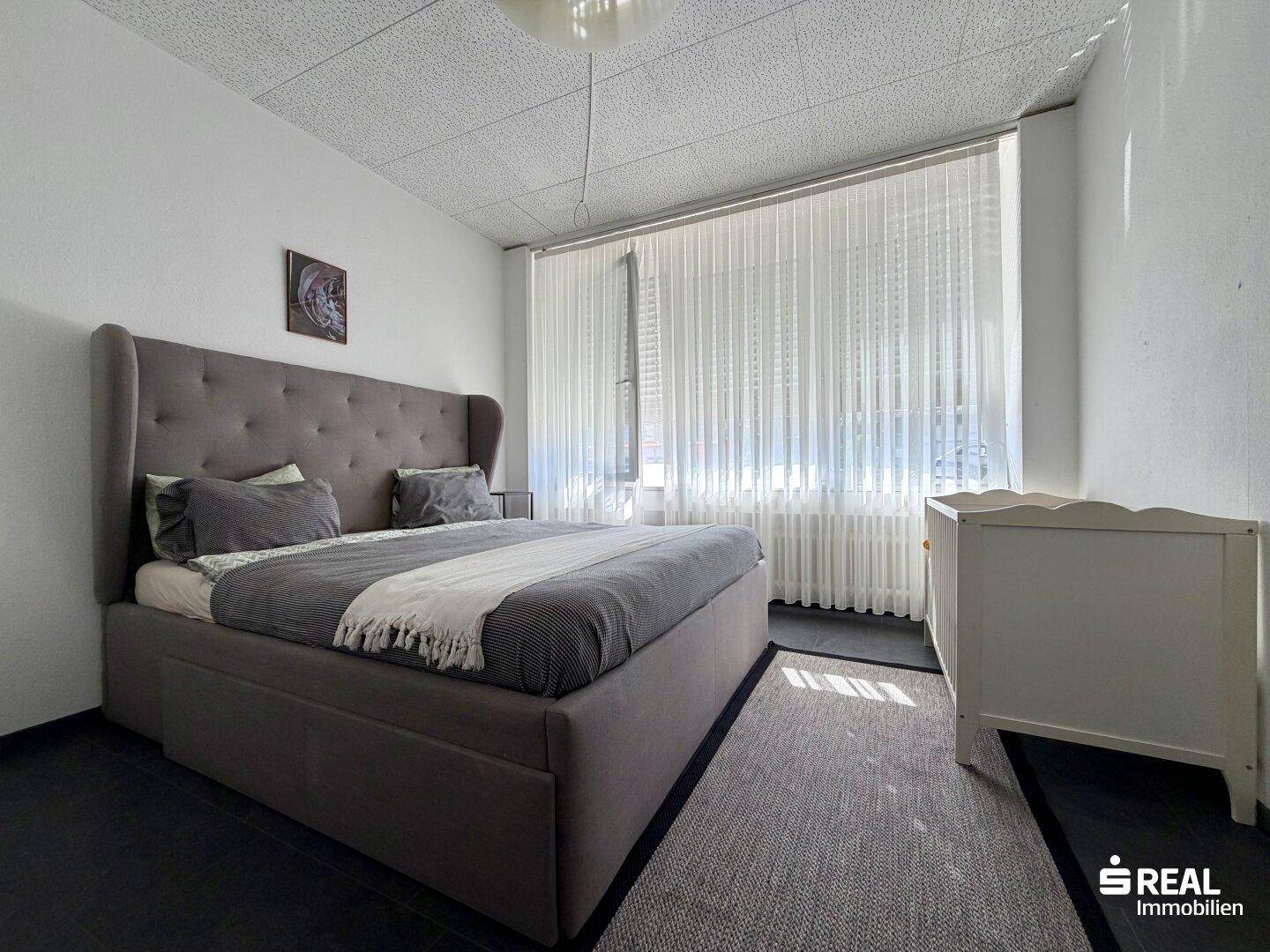 Helles Schlafzimmer mit großem Polsterbett, Babybett und viel Tageslicht durch große Fenster.