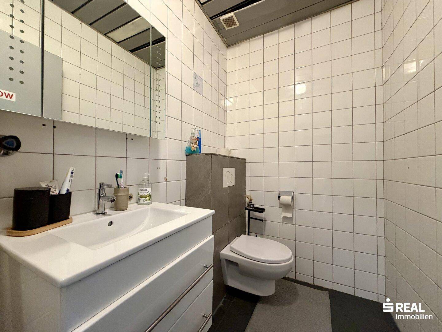 Badezimmer mit weißen Fliesen, WC, Waschtisch und Spiegelschrank.