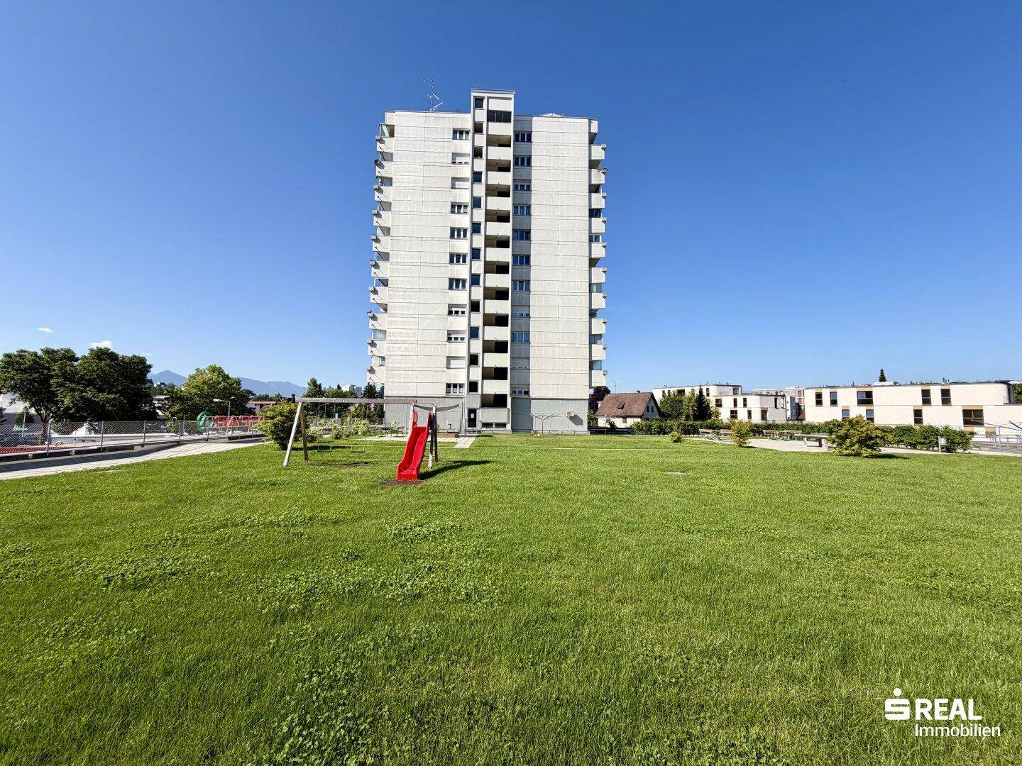 Hohes Mehrfamilienhaus mit großer Grünfläche und Kinderspielplatz im Vordergrund.