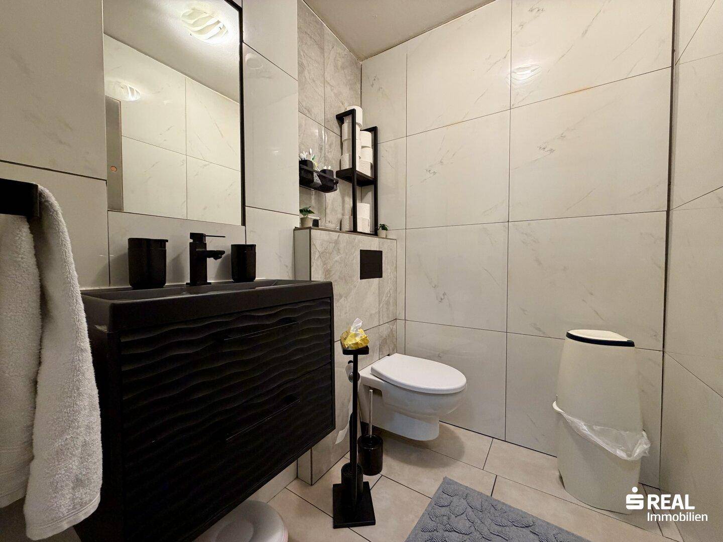 Modernes WC mit wandmontierter Toilette, schwarzem Waschbecken und hellen Marmorfliesen.